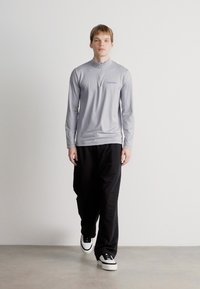 Γκρι μπλουζάκι μακρύ με μιμούμενο γιακά, συνδυασμένο με χαλαρές μαύρες παντελόνες. Το μοντέλο φοράει λευκά αθλητικά παπούτσια με μαύρες λεπτομέρειες σε ουδέτερο φόντο.