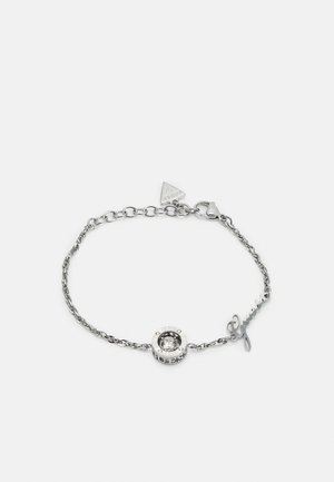 Pulseira de corrente prateada com um charme em forma circular, uma pedra transparente, acentos gravados e um detalhe em script "Guess".