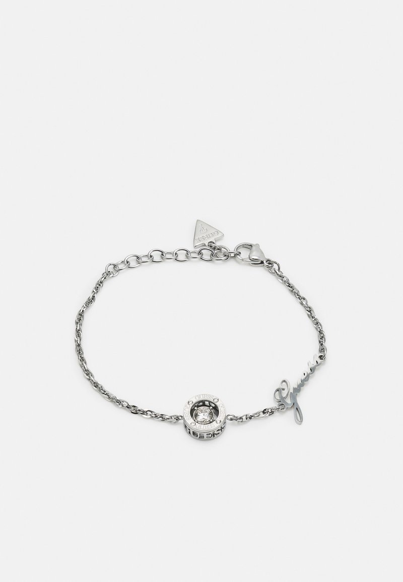 Bracelet en chaîne argentée avec un charme circulaire orné d'une pierre transparente, des accents gravés et un détail en écriture "Guess".
