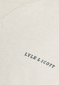 Κοντινό πλάνο ανοιχτόχρωμο υ μπεζ ύφασματος με μαύρο κεντημένο κείμενο "LYLE & SCOTT" κοντά σε ριμπλ γραμμή λαιμού.