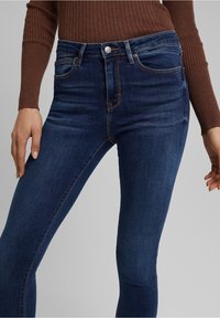 Jeans ajustados en denim azul oscuro, con una cintura de altura media, cinco bolsillos y sutiles acentos de costura en naranja. Textura suave, diseño entallado.