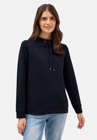 Mörkblå sweatshirt med hög krage, snörning, långa ärmar, ribbade mudd och en mjuk textur, kombinerad med ljusblå jeans.