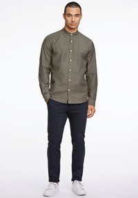 Camisa de manga larga verde oliva con cuello mao, cierre de botones y fabricada en algodón, combinada con pantalones ajustados azul marino y zapatillas blancas.