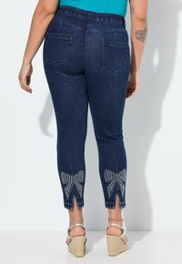 Tmavé denimové tapered džíny s vysokým pasem. Obsahují vykrojený lem zdobený kamínkovými mašličkami, pět kapes a tónové švy.