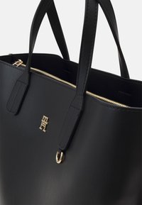 Zwarte leren tote bag met gouden hardware, dubbele handvatten en een ritssluiting. Beschikt over een gestructureerd oppervlak en een prominent logo aan de voorkant.