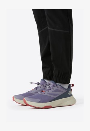 Chaussures de sport violettes avec une tige texturée, des lacets blancs, des accents rouges et une semelle beige. Portées avec un pantalon noir fuselé.