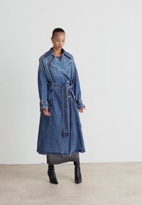 Denim trenchcoat, lang, dubbelzijdig met brede kraag, ceintuur in de taille en opgerolde manchetten. Donkerblauwe kleur, licht vervaagde textuur.