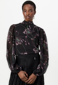Blusa nera trasparente con stampa floreale in viola e verde, collo alto, maniche a sbuffo e polsini arricciati. Abbinata a una gonna nera plissettata.