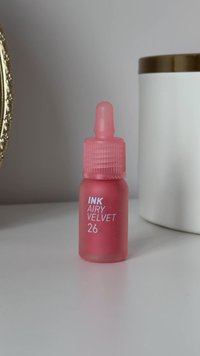 Bottiglia con contagocce in vetro rosa con cappuccio rigato, etichettata "INK AIRY VELVET 26," posizionata su una superficie bianca accanto a un contenitore cilindrico bianco.