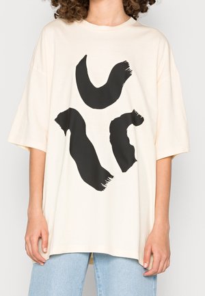 Personne portant un t-shirt crème ample avec trois formes abstraites en coups de pinceau noirs à l'avant, associé à un jean bleu clair.