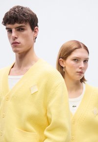 Κίτρινες πλεκτές ζακέτες με V-λαιμόκοψη, κουμπιά μπροστά και τσέπη. Μία έχει σχέδιο με διαμάντι. Φοριούνται πάνω από λευκά τοπ. Μινιμαλιστικός σχεδιασμός.