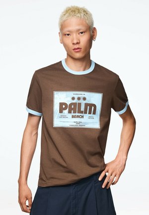 Mladá osoba s krátkymi blond vlasmi, ktorá má na sebe hnedé tričko so svetlomodrým lemom a textovou grafikou "Palm Beach", stojaca na jednoduchom bielom pozadí.