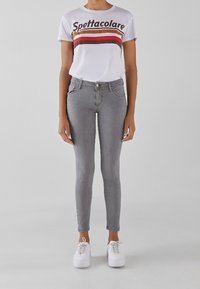 Jeans ajustados grises combinados con una camiseta blanca que presenta un estampado gráfico de rayas rojas y naranjas. El modelo lleva zapatillas blancas.