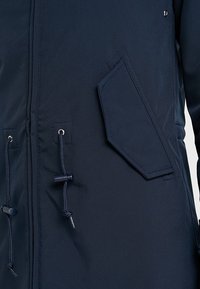 Mørkeblå jakke med en glat tekstur, der har en skrå lomme-design og snørebånd i taljen og kraven for justerbar pasform.