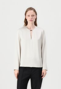 Licht beige blouse met lange mouwen en een sleutelhole halslijn, gladde textuur en minimalistisch ontwerp. Gecombineerd met zwarte broeken.