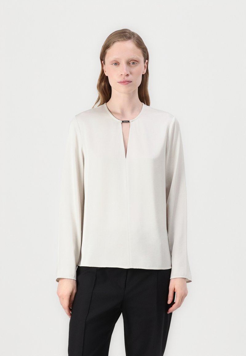 Licht beige blouse met lange mouwen en een sleutelhole halslijn, gladde textuur en minimalistisch ontwerp. Gecombineerd met zwarte broeken.