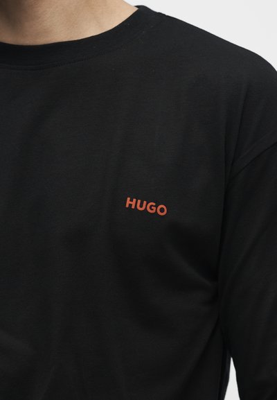 Chemise noire à manches longues avec un petit logo orange "HUGO" sur la poitrine gauche, portée par une personne visible du cou jusqu'au milieu du torse.