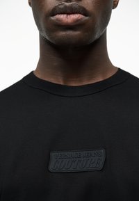 Κοντινό πλάνο ενός ατόμου που φοράει μαύρο t-shirt Versace Jeans Couture, εμφανίζοντας το κάτω μέρος του προσώπου, τον λαιμό και το πάνω μέρος του στήθους.