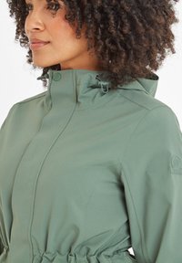 Green softshell jacket featuring a high collar, adjustable drawstring, and fitted waist. Textured fabric with visible seams and logo detail.