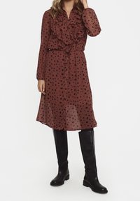 Brązowa przezroczysta sukienka w czarne kropki, z falbanami na kołnierzu i z przodu, z zapięciem na guziki i długimi r�ękawami. Uzupełniona czarnymi, wysokimi do kolan botkami.