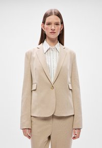 Béžový štruktúrovaný blazer s priliehavým strihom, klopami, dvoma prednými vreckami a uzatváraním na jeden gombík. Nosený cez bielu pruhovanú košeľu.