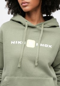 Olivovo zelená mikina s kapucňou, vybavená predným vreckom typu klokanie vrecko, sťahovacou šnúrkou na kapucni a bielym potlačami "NIKE" a "SHOX" v svetlom zvýrazňujúcom ráme.