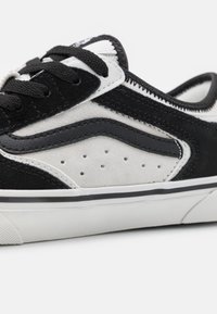 Zapatilla de ante negra y gris con una superficie texturizada, que presenta costuras blancas, cordones negros y una suela de goma blanca con una franja negra.