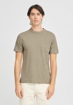 T-shirt khaki a maniche corte in un morbido tessuto testurizzato con collo a giro e una tasca sul petto. Indossato con pantaloni chiari.
