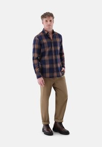 Karierte Hemdjacke in Marineblau, Orange und Creme; beigefarbene Hose; braune Schnürstiefel; Modell steht mit einer Hand in der Tasche.