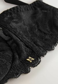 Zwart kant stof met scalloped randen, met een klein gouden logo accent. Zachte textuur, gedetailleerd patroon zichtbaar over de hele stof.
