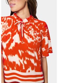 Blusa rossa a fantasia con scollatura arricciata e dettaglio con fiocco. Maniche corte, caratterizzata da un design a righe orizzontali all'orlo. Tessuto liscio.