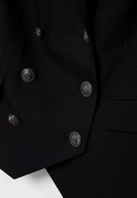 Gros plan d'un blazer noir croisé à six boutons décoratifs argentés et une patte de poche avant côté droit.