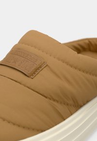 Rudos spalvos slip-on bateliai su minkštu tekstūru, ant jų matomas tekstūruotas prekės ženklo ženklas ir balta vidurinė pado dalis. Dizainas yra minimalus ir elegantiško silueto.