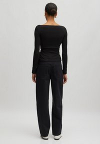 Schwarzes Langarmoberteil mit quadratischem Ausschnitt, kombiniert mit hochgeschnittenen, weit geschnittenen schwarzen Jeans und weißen Schuhen, von hinten gesehen.