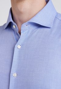 Close-up van een persoon in een lichtblauw gestructureerd overhemd met witte knopen, waarbij de kraag en het bovenste deel van de borst zichtbaar zijn.