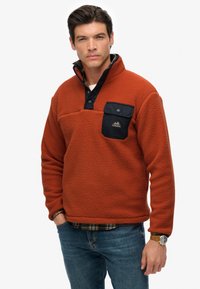 Orange fleece pullover with a textured finish, black buttoned collar and chest pocket, paired with blue jeans and a watch with a brown strap.