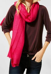 Jasnoróżowy szal z miękkiego materiału, luźno zarzucony na szyi, z frędzlami na krawędzi. W połączeniu z ciemnobrązową bluzką i dżinsami.
