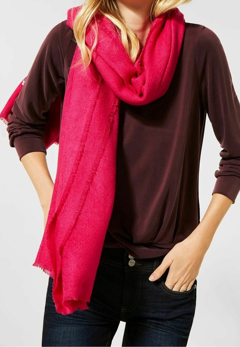 Jasnoróżowy szal z miękkiego materiału, luźno zarzucony na szyi, z frędzlami na krawędzi. W połączeniu z ciemnobrązową bluzką i dżinsami.