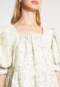 Femme portant une robe blanche texturée avec des manches bouffantes et un décolleté carré, accessoirisée de grandes boucles d'oreilles dorées.