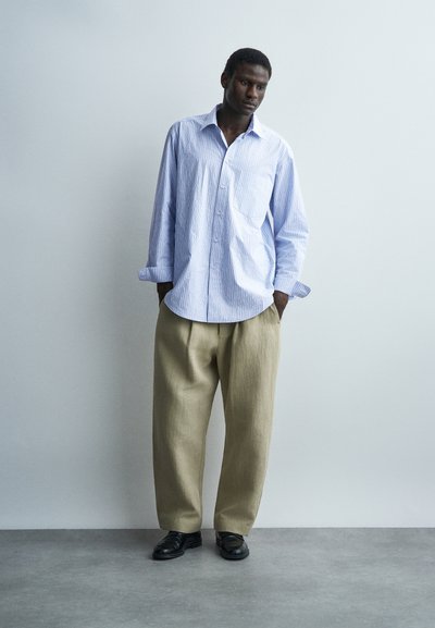 Άνδρας που στέκεται με τα χέρια στις τσέπες, φοράει ανοιχτό μπλε ριγέ πουκάμισο σε υπερμεγέθη γραμμή, μπεζ παντελόνι με φαρδιά πόδια και μαύρα παπούτσια, σε απλό φόντο.
