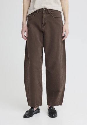 Person iført løstsiddende brune bukser og sorte loafers, stående mod en enkel lys baggrund.