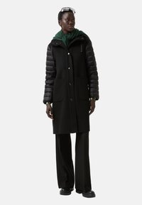 Sort frakke med puffede sorte ærmer, to forlommer og et grønt tørklæde. Designet har trykknapper og en skræddersyet form.