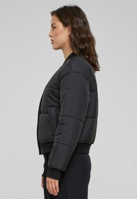 Černá péřová bomber bunda s žebrovanými manžetami, kulatým límcem a bočními kapsami, s hladkým matným povrchem a hranatým střihem.