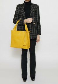 Gele leren tote bag met twee handgrepen, rechthoekige vorm, gladde textuur, met een trekkoordsluiting en een klein etiketdetail.