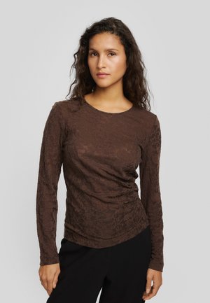 Une femme aux longs cheveux foncés pose devant un fond gris uni. Elle porte un haut marron à manches longues et à motifs, ainsi qu’un pantalon noir.
