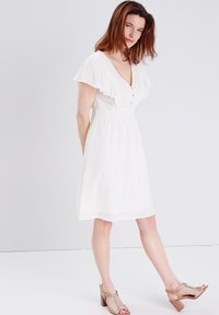 Weißes Kleid mit tiefem V-Ausschnitt und rüschigen kurzen Ärmeln. Verfügt über Knopfakzente und einen strukturierten Stoff. Kombiniert mit beigen, rutschfesten Absätzen.