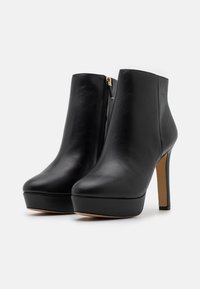 Botines de cuero negro con acabado brillante, con base de plataforma y tacón alto de aguja. Cierre lateral con cremallera; textura lisa.