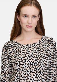 Schwarze und cremefarbene Bluse mit Muster, mit Falten-Details und rundem Ausschnitt. Der Stoff hat eine glatte Textur und zeigt abstrakte Formen.