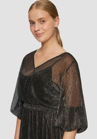 Schwarzes Kleid mit durchscheinenden, strukturierten Ärmeln und tiefem V-Ausschnitt. Mit metallischen Fäden für Glanz, tailliert und mit fließendem Rock.