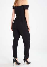 Kvinna iklädd en figurnära, off-shoulder svart jumpsuit med avsmalnande ben och svarta högklackade skor, ståendes vänd bort, mot vit bakgrund.
